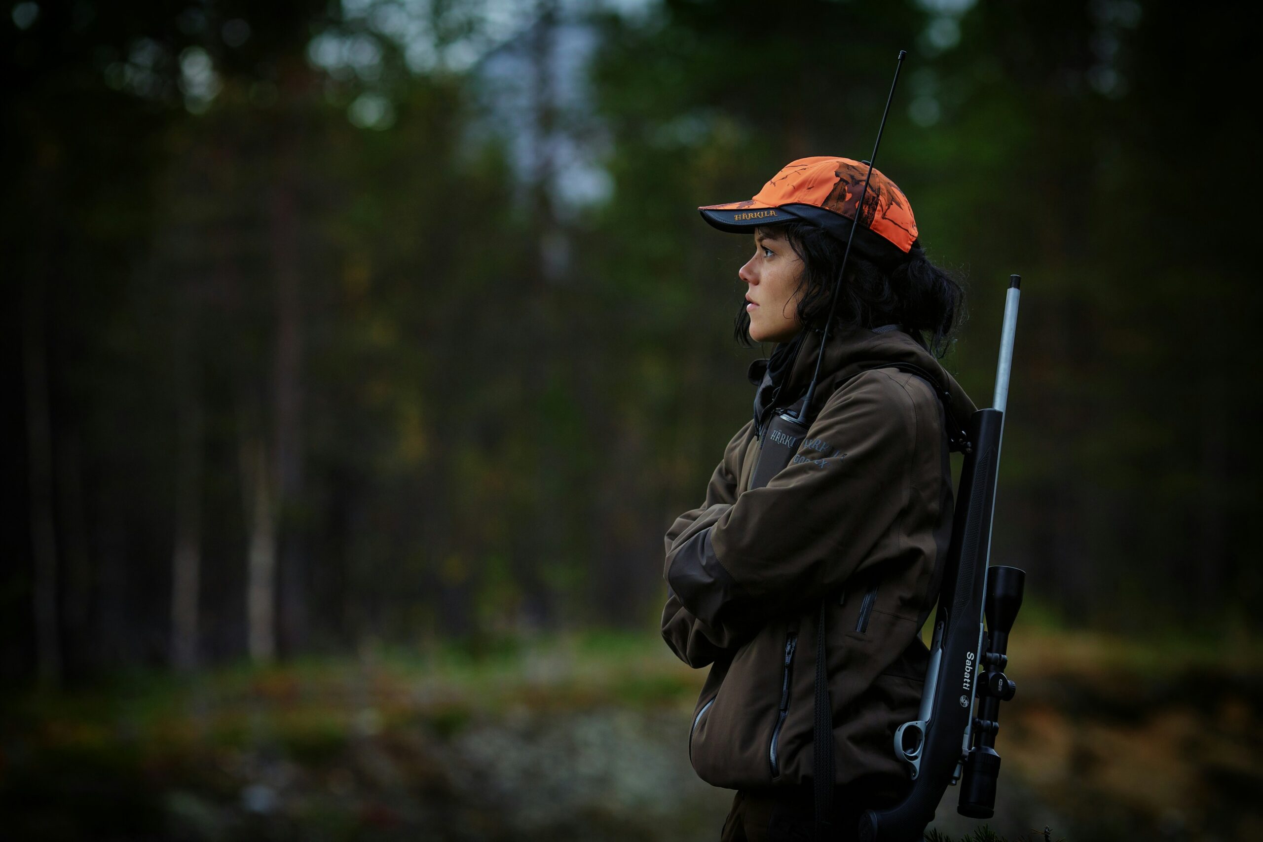Hunter in blaze orange cap carrying a scoped rifle in a forested hunting area